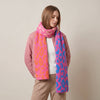A woman wearing a fuzzy knit scarf with a pink, orange, and blue leopard print pattern, styled with a pink jacket and beige pants against a neutral beige background.