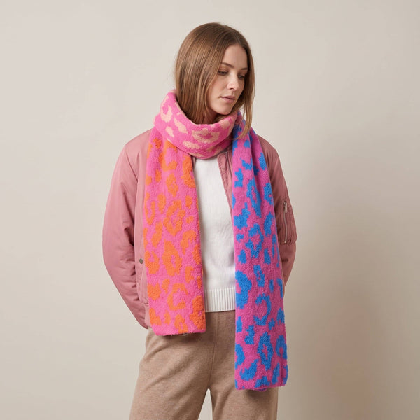 A woman wearing a fuzzy knit scarf with a pink, orange, and blue leopard print pattern, styled with a pink jacket and beige pants against a neutral beige background.