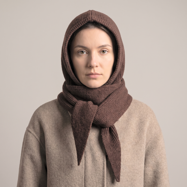 A woman wearing a brown ribbed knit hood and matching scarf over a beige coat, photographed against a plain light gray studio background.