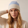 A woman wearing a ribbed knit beanie hat with a light blue top section and a cream brim, paired with a cream knit sweater against a plain light background.